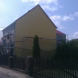 Żółty dom jednorodzinny w trakcie prac elewacyjnych, widoczne rusztowania i pracownicy, ogrodzenie z kamiennymi słupkami na pierwszym planie.