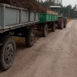 Ciągnik rolniczy z dwiema przyczepami wypełnionymi gałęziami i korzeniami, stojący na polnej drodze w pochmurny dzień. W tle widoczne drzewa.