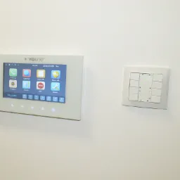 Panel sterowania domofonem marki Vidiline z ekranem dotykowym i przyciskami funkcyjnymi, zamontowany na białej ścianie, obok panel z włącznikami światła.