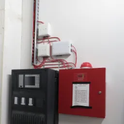 Panel sterowania systemem przeciwpożarowym z czerwonymi przewodami, umieszczony na białej ścianie obok czerwonej skrzynki z instrukcją.