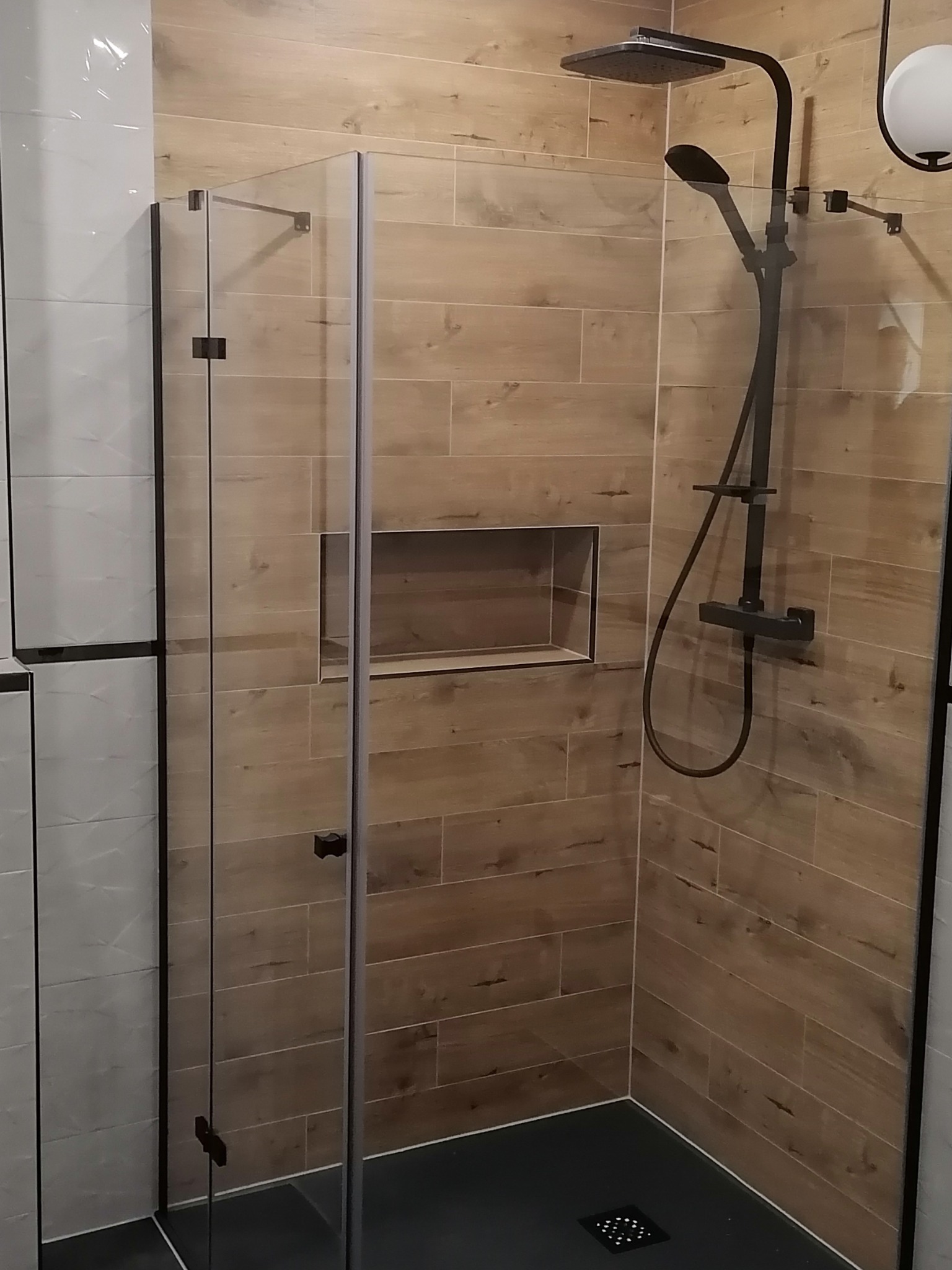 Kabina prysznicowa z czarną armaturą i drewnopodobnymi płytkami na ścianie, wbudowana półka w ścianie, czarny brodzik.