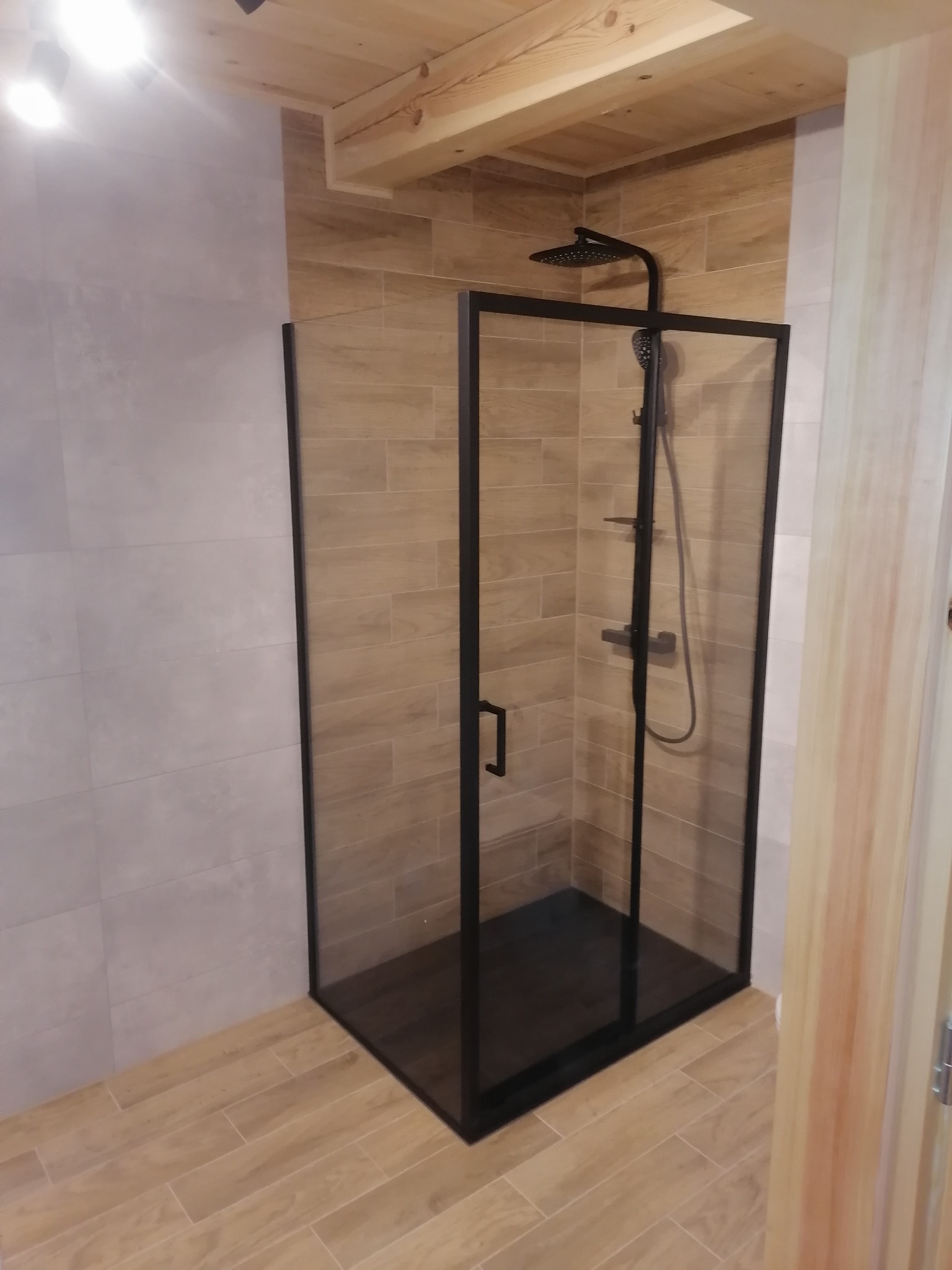 Kabina prysznicowa z czarnymi ramami w łazience wykończonej płytkami imitującymi drewno i szarymi płytkami, z drewnianym sufitem.