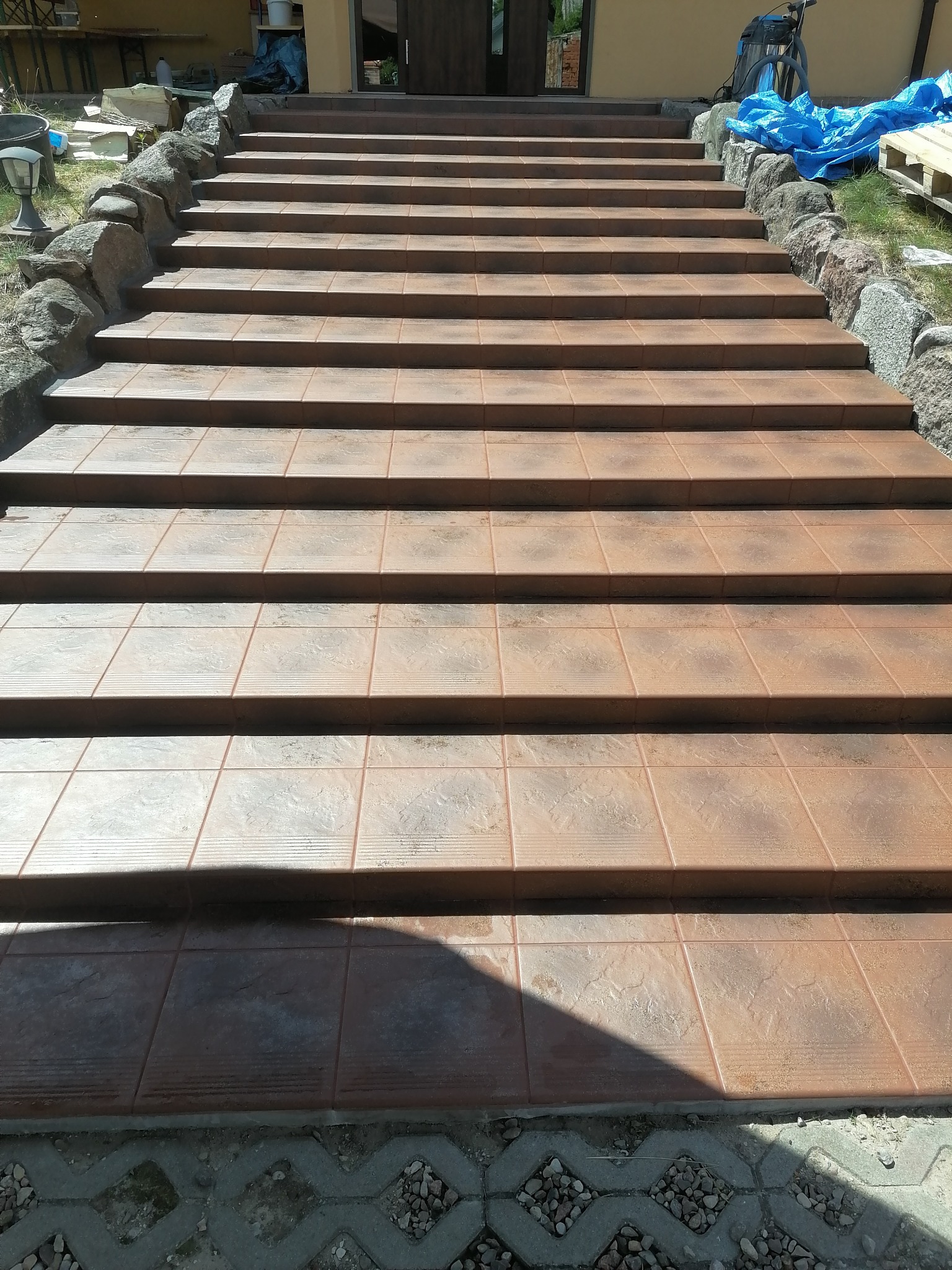 Zewnętrzne schody wyłożone brązowymi płytkami z dekoracyjną fakturą, otoczone kamieniami, prowadzące do drzwi wejściowych.