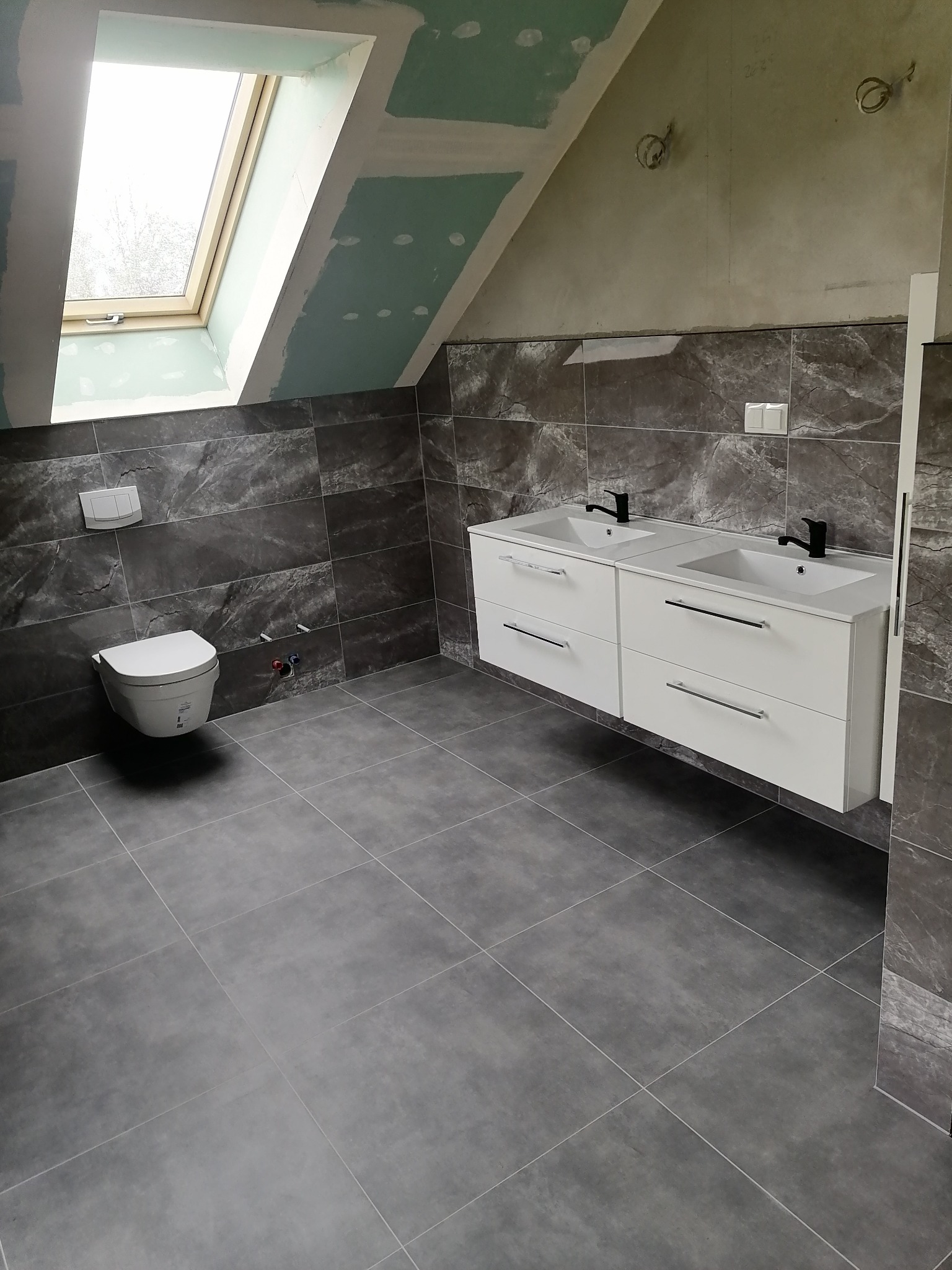 Ukończona łazienka na poddaszu z szarą glazurą imitującą marmur, wiszącą toaletą i podwójną białą szafką z umywalkami i czarnymi bateriami, widoczne niedokończone gładzie na suficie pod oknem...