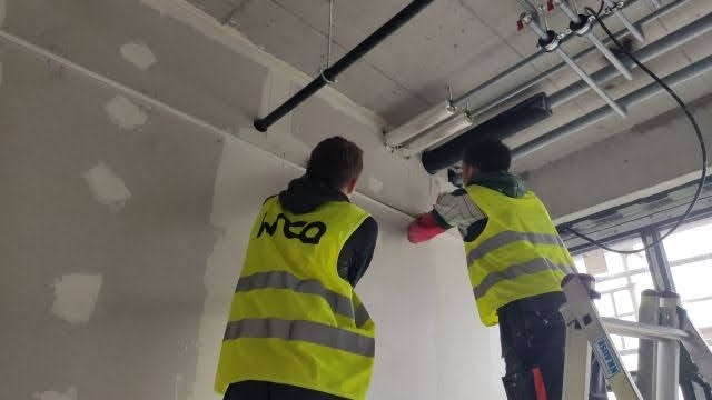 Dwóch pracowników w kamizelkach, na drabinie, szpachluje ścianę z widocznymi łatami gipsowymi. W tle instalacje pod sufitem.