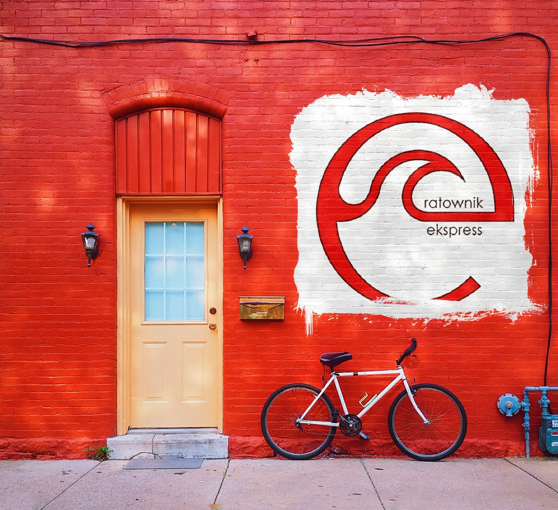 Czerwona ceglana ściana z beżowymi drzwiami, stylizowanym logo 'ratownik ekspress' i białym rowerem opartym o ścianę