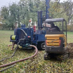 Wiertnica na trawiastym terenie z wykopem i stertą ziemi obok. Widoczny operator w kabinie minikoparki Volvo. W tle drzewa i pochmurne niebo.