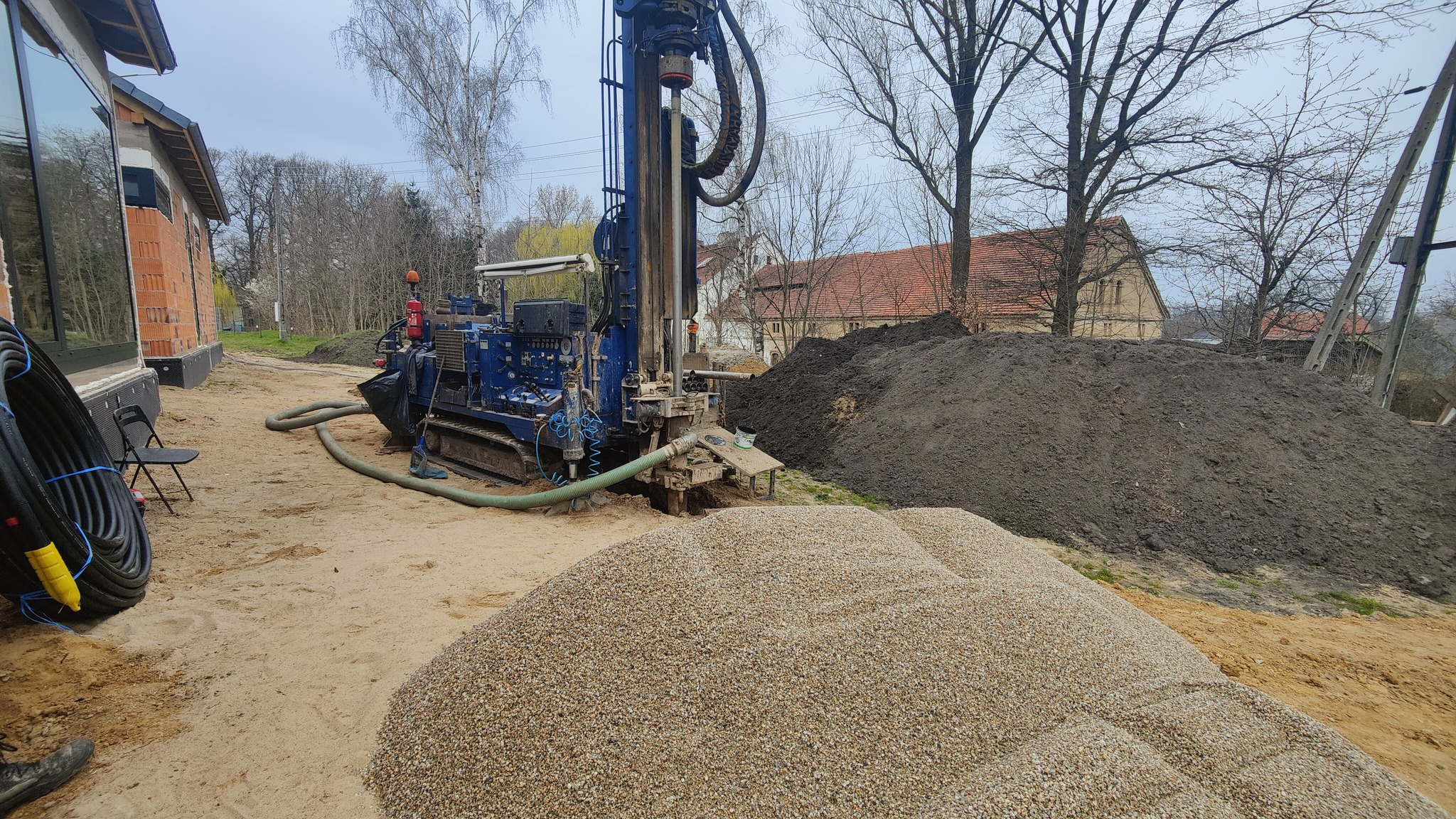 Wiertnica na gąsienicach podczas pracy przy budowie studni głębinowej, obok hałda ziemi i żwiru. W tle budynek z czerwonej cegły i drzewa.