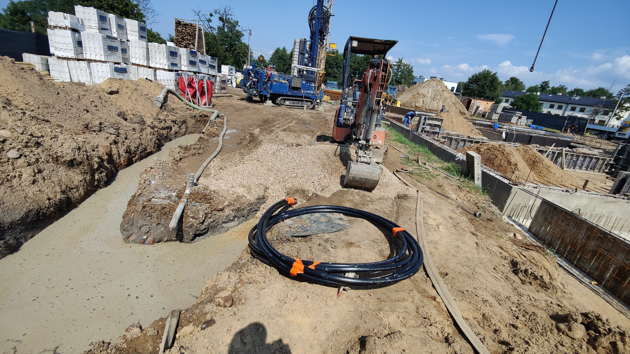 Wykop z rurami i sprzętem do wiercenia studni głębinowej. Widoczny wykop, rury zwinięte w krąg, koparka i wiertnica na placu budowy w słoneczny dzień.