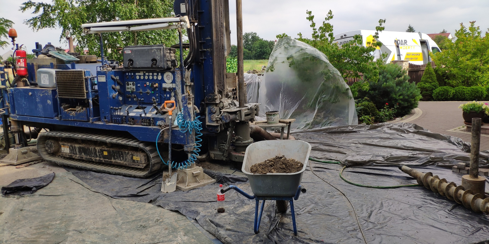 Mobilna wiertnica do studni głębinowych na gąsienicach, z wózkiem pełnym ziemi i świdrem w tle. Widoczny samochód dostawczy firmy. Teren zabezpieczony folią.