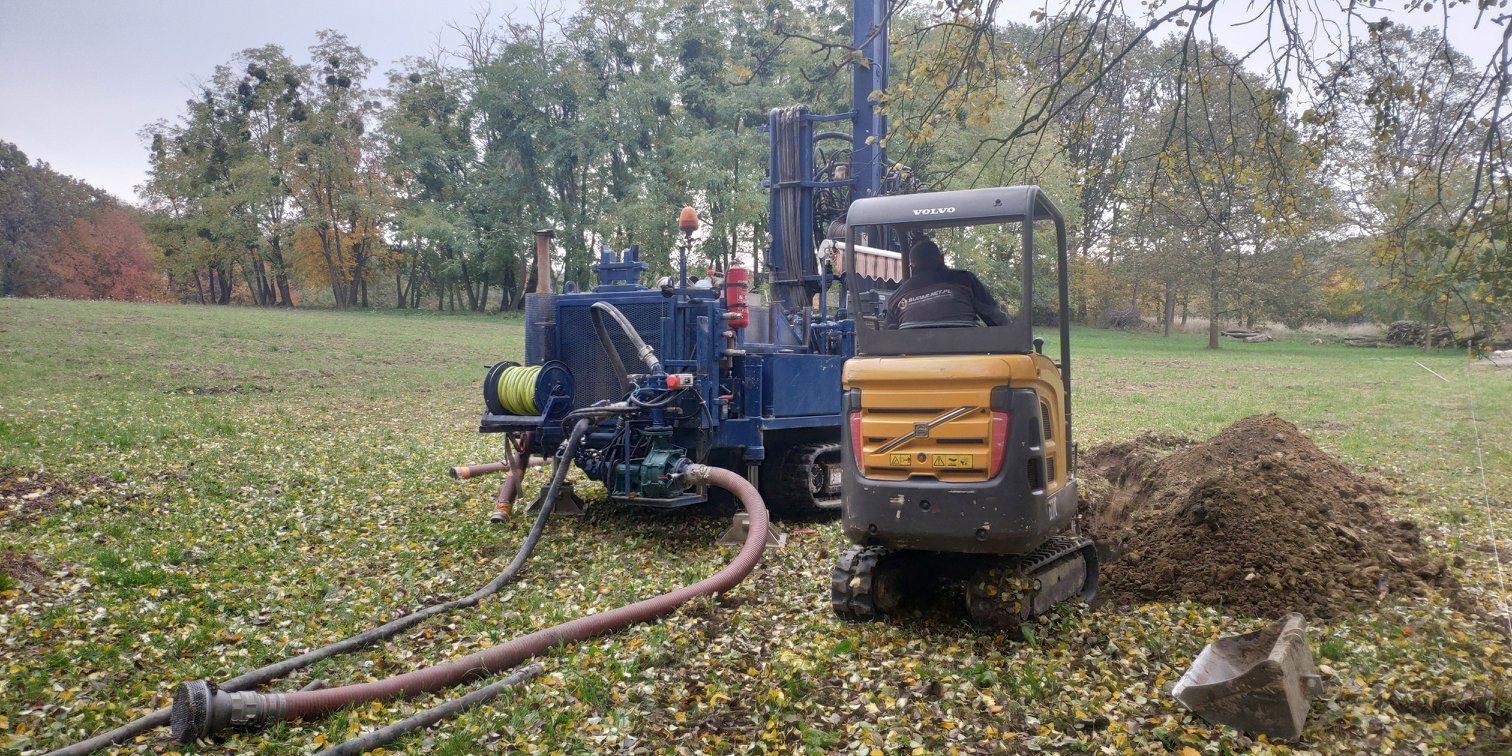 Wiertnica na trawiastym terenie z wykopem i stertą ziemi obok. Widoczny operator w kabinie minikoparki Volvo. W tle drzewa i pochmurne niebo.