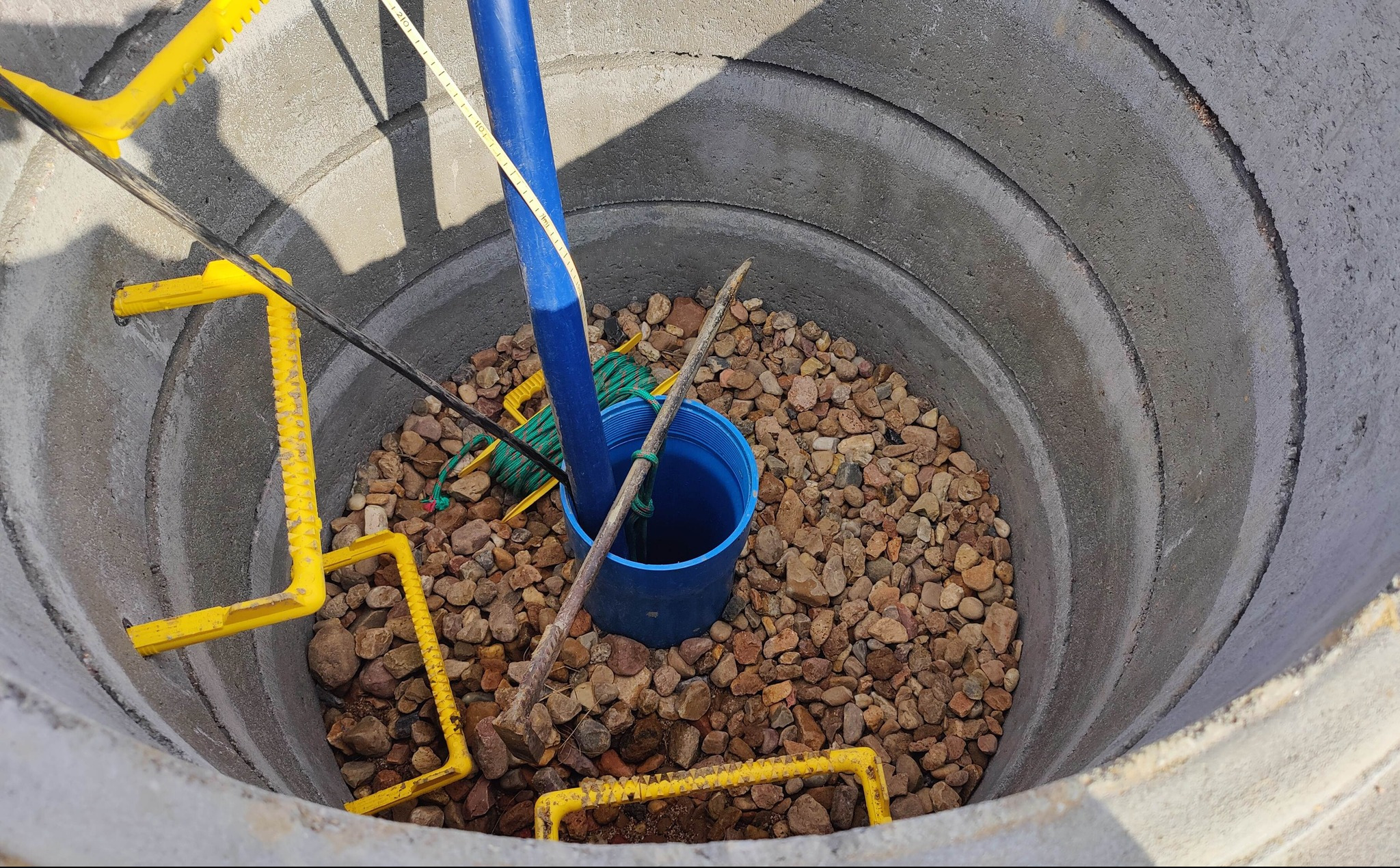 Wnętrze betonowej studni kręgowej wypełnionej żwirem, z niebieską rurą i elementami żółtej drabinki. Widać też zielony sznur i niebieski pojemnik.