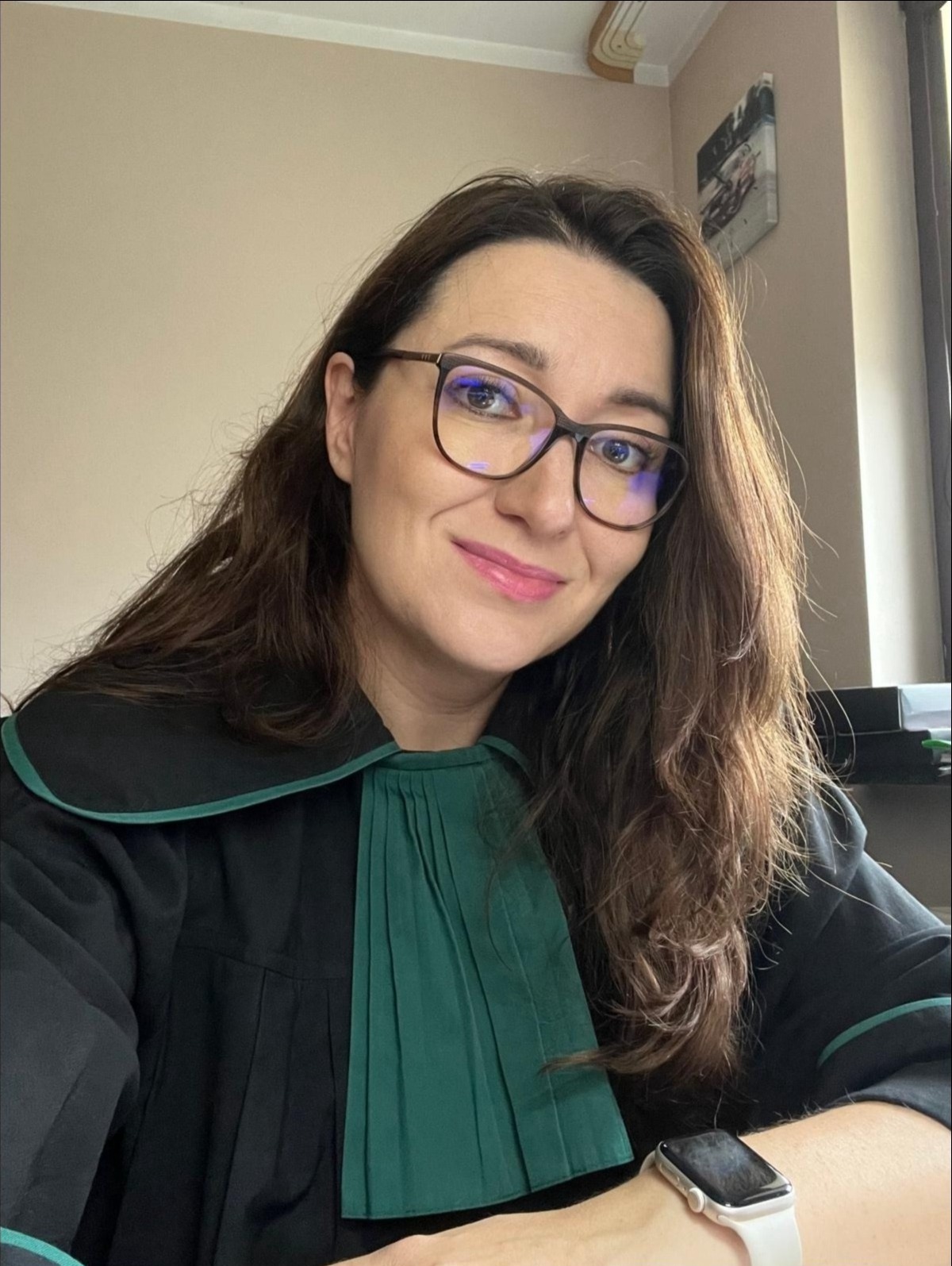 Uśmiechnięta kobieta w todze adwokackiej, z brązowymi włosami i okularami, siedząca przy biurku w jasnym pomieszczeniu. Widoczny zegarek na nadgarstku.