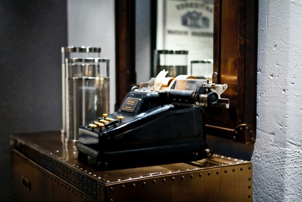 Czarna, zabytkowa maszyna licząca z żółtymi przyciskami, umieszczona na metalowej komodzie w biurze w Gorzycach, obok szklanych pojemników i lustra z drewnianą ramą.