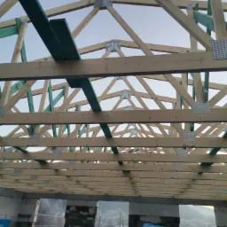 Konstrukcja drewniana dachu w trakcie budowy, widoczne elementy więźby dachowej z metalowymi łącznikami, ściany z pustak&oacute;w oraz materiały budowlane Porfix