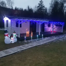 Biały budynek z drewnianą elewacją, oświetlony niebieskimi lampkami, z dekoracjami świątecznymi: dmuchane bałwany, renifery i gwiazdki. Kamienna ścieżka prowadzi do wejścia.