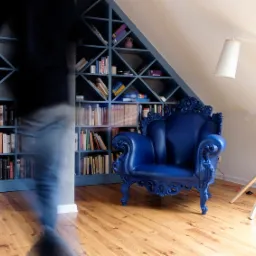 Niebieski, bogato zdobiony fotel na drewnianej podłodze, na tle niebieskiego regału z książkami, obok biała lampa stojąca, zamazana sylwetka osoby w ruchu.
