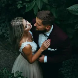 Para młoda w objęciach, panna młoda w koronkowej sukni z odkrytymi ramionami, pan młody w bordowym garniturze i muszce, otoczeni zielenią, ujęcie z góry.