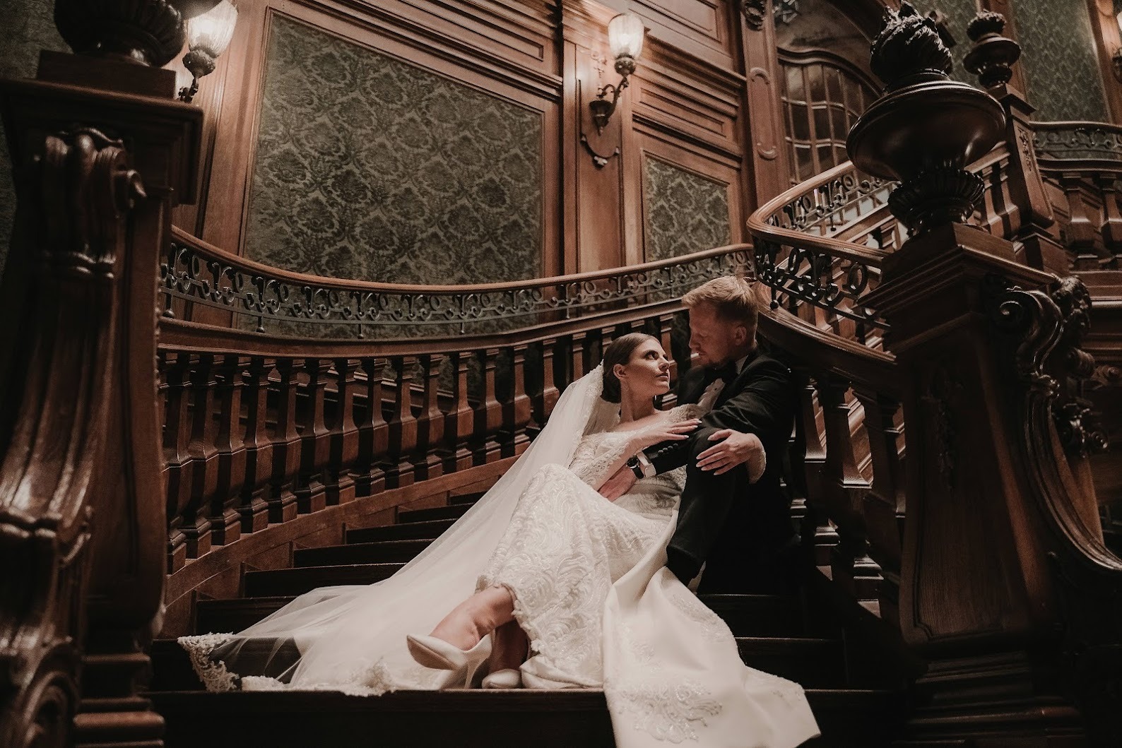 Para młoda siedzi na drewnianych schodach w eleganckim wnętrzu, panna młoda w koronkowej sukni i białych butach, pan młody w czarnym smokingu, spoglądają na siebie z czułością.