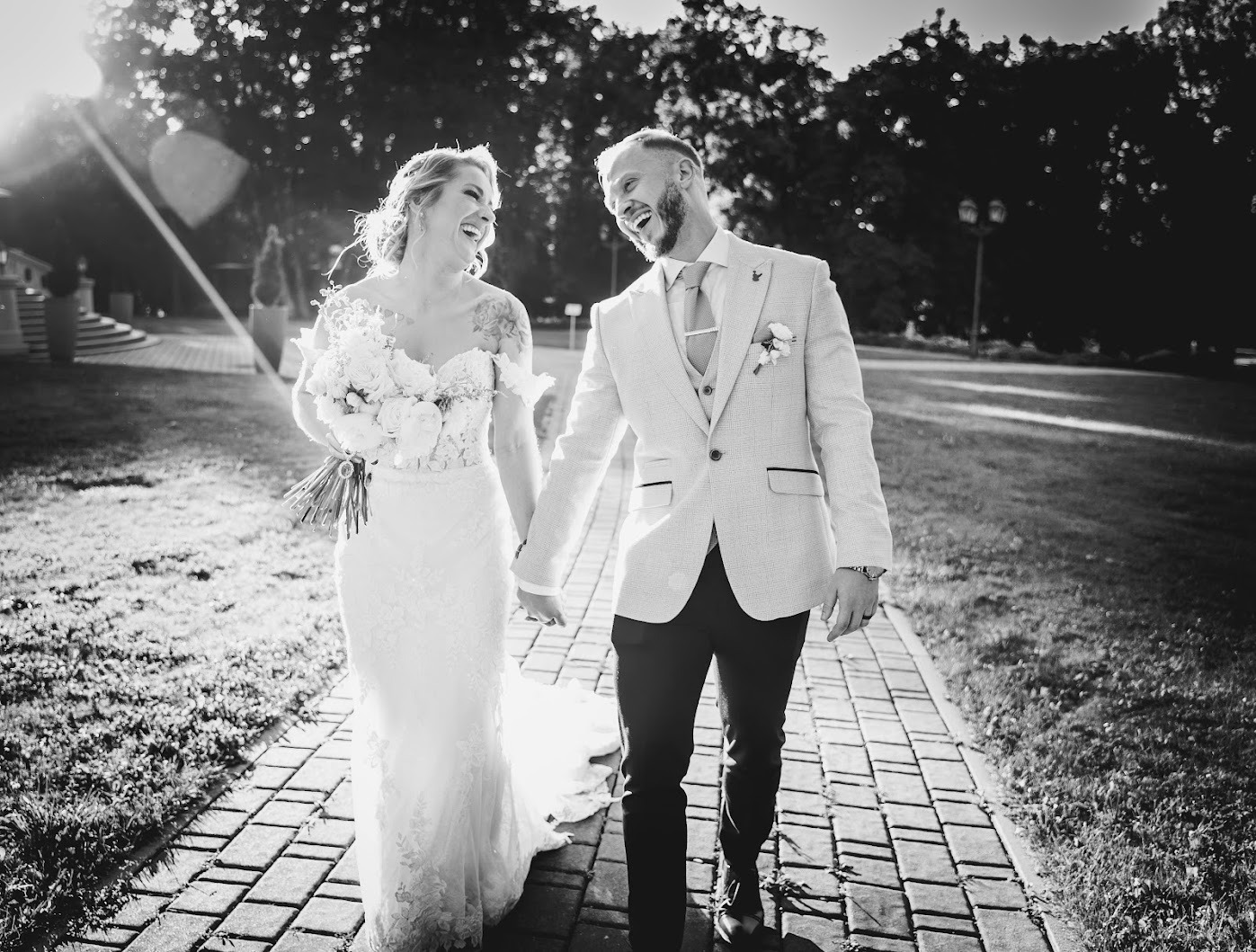 Czarno-białe zdjęcie uśmiechniętej pary młodej, spacerującej po brukowanej ścieżce w Katowicach, panna młoda w koronkowej sukni trzyma bukiet, pan młody w jasnym garniturze, w tle drzewa i budynek.