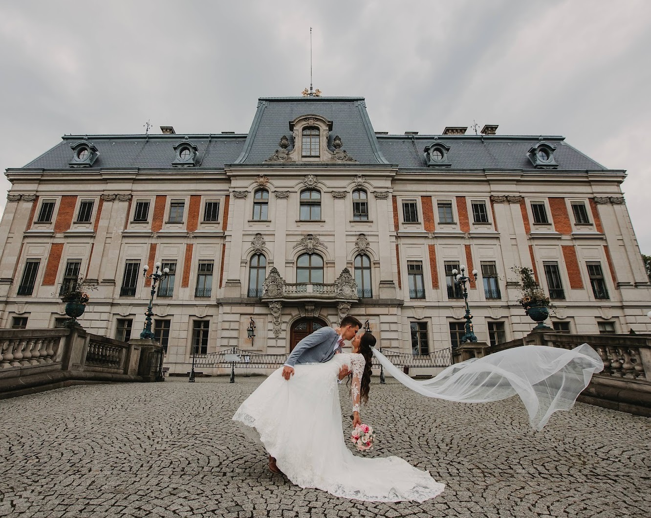 Pocałunek nowożeńców na tle pałacu, welon powiewa na wietrze, brukowana droga.