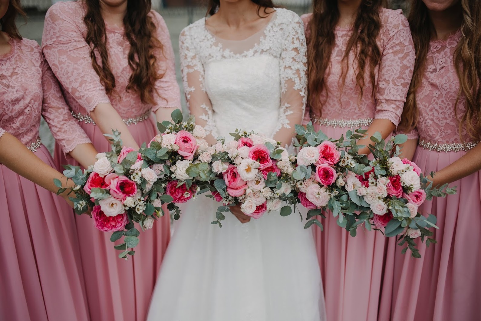 Panna młoda w białej sukni i druhny w różowych koronkowych sukienkach trzymają wspólnie bukiet różowych i białych róż z eukaliptusem.