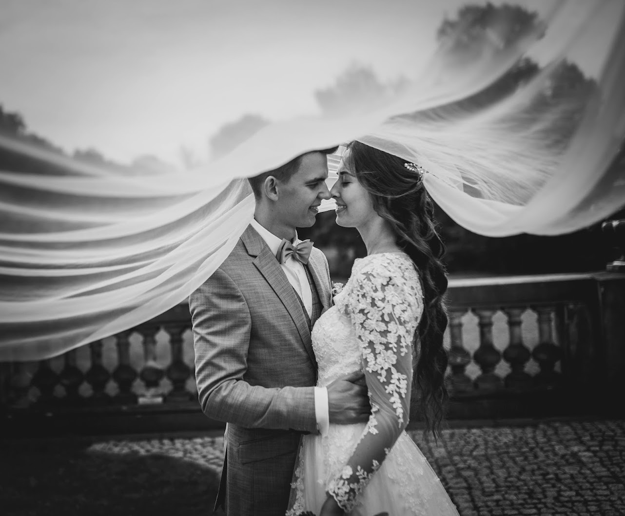 Czarno-białe zdjęcie nowożeńców pod welonem, delikatnie się dotykających nosami, mężczyzna w szarym garniturze w kratę, kobieta w koronkowej sukni z długim rękawem, włosy upięte z boku.