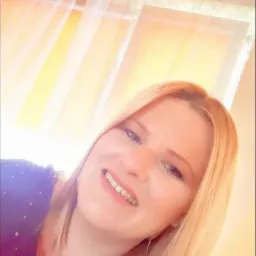 Uśmiechnięta blond kobieta w ciemnej bluzce z błyszczącymi aplikacjami, z okrągłymi kolczykami, pozująca na tle okna z białymi firankami.