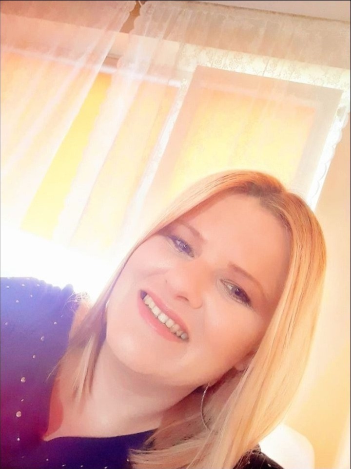 Uśmiechnięta blond kobieta w ciemnej bluzce z błyszczącymi aplikacjami, z okrągłymi kolczykami, pozująca na tle okna z białymi firankami.