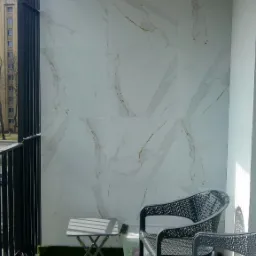Balkon z wyłożoną ścianą płytkami imitującymi marmur, sztuczna trawa na podłodze, dwa czarne krzesła z ażurowym wzorem i mały stolik.