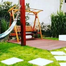 Drewniana huśtawka ogrodowa z hamakiem, ustawiona na tarasie z desek kompozytowych, otoczona zielenią i ścieżką z płyt chodnikowych.