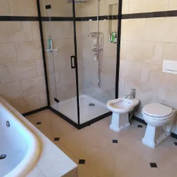 Wykończona łazienka z kabiną prysznicową typu walk-in z czarną ramą, białą wanną, bidetem i toaletą, wyłożona beżowymi płytkami z czarnymi akcentami na podłodze i paskiem dekoracyjnym na ścianie.