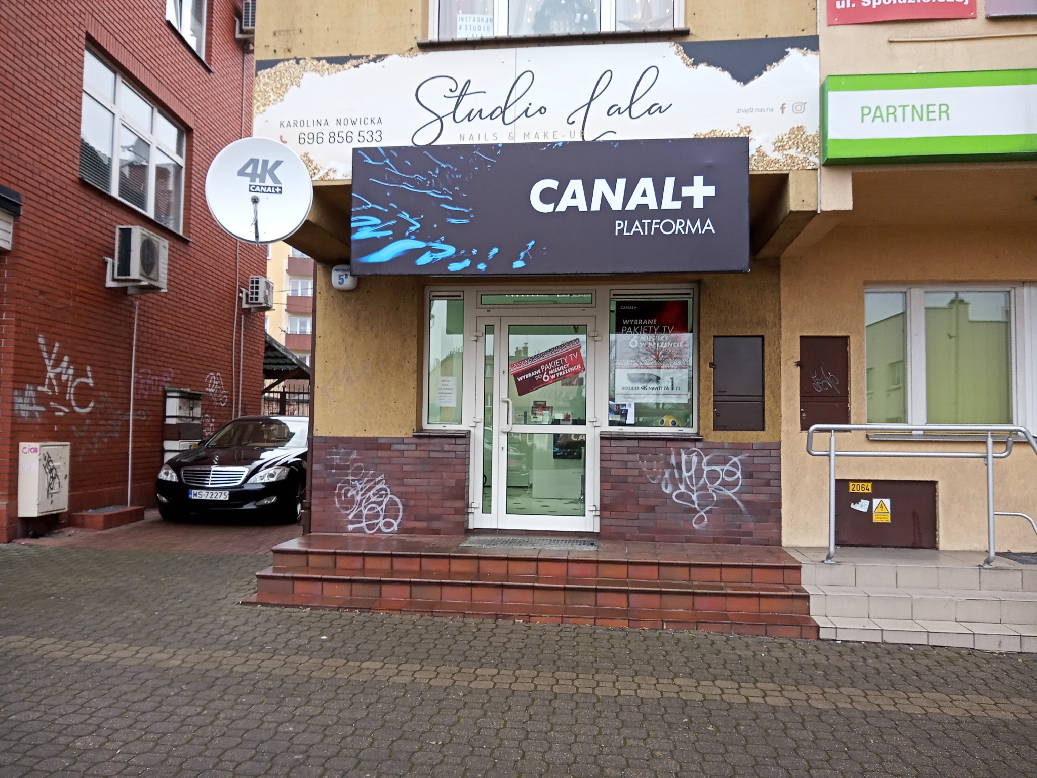 Wejście do punktu sprzedaży Canal+ Platforma z szyldem nad drzwiami i anteną satelitarną 4K na sąsiednim budynku z czerwonej cegły.