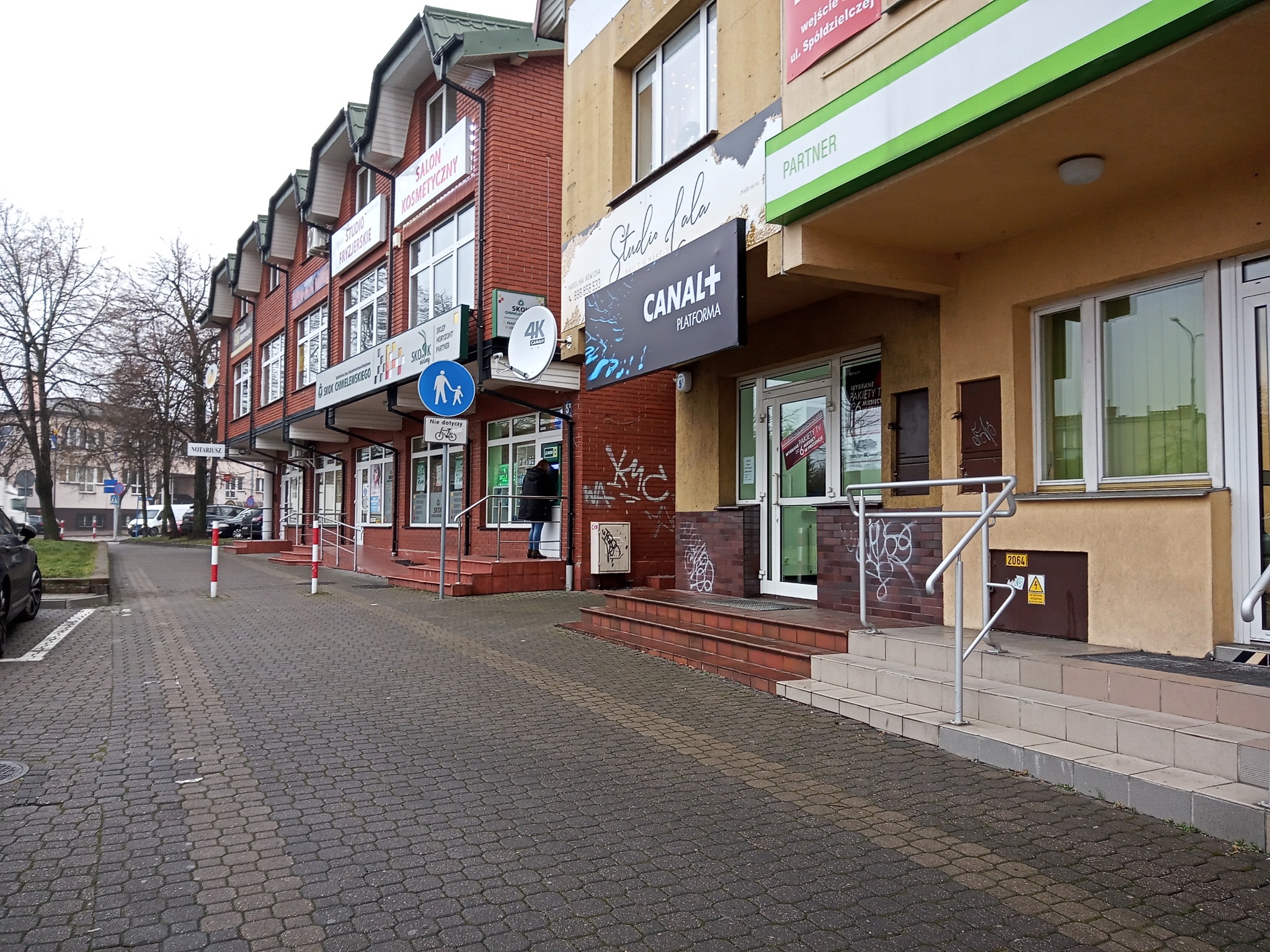 Fasady budynków usługowych w Łukowie, z szyldami reklamowymi Canal+ i Studio Urody, widoczne z chodnika wyłożonego kostką brukową.
