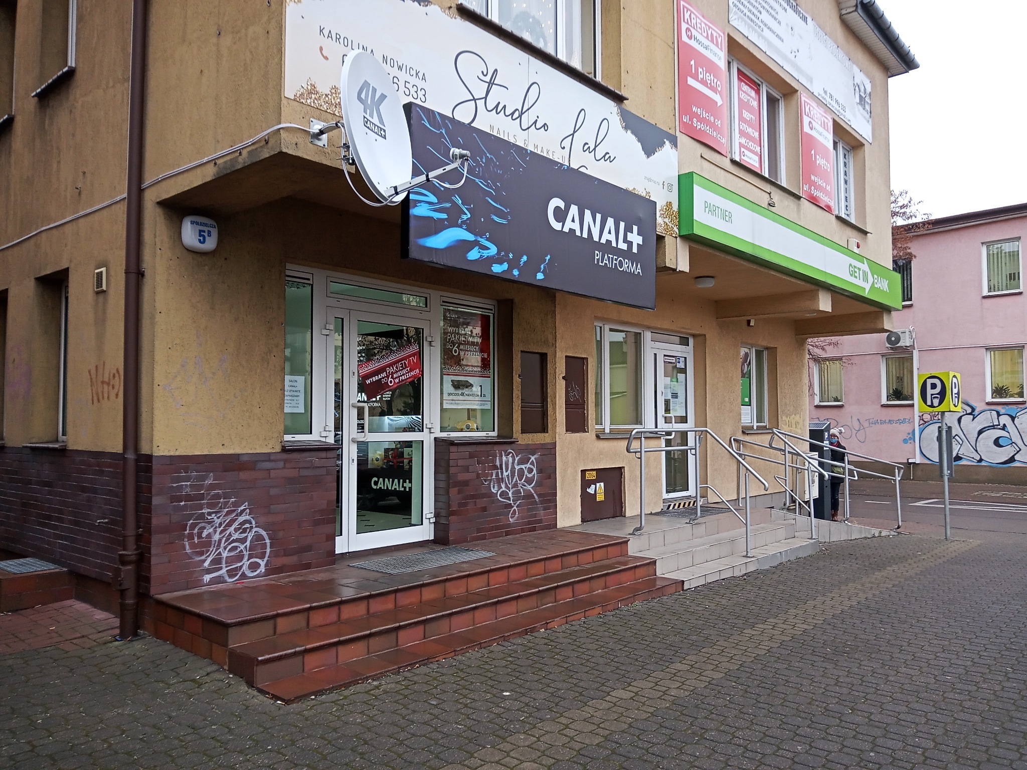 Fasada budynku z szyldami reklamowymi, w tym Canal+ Platforma i Studio Fala, widoczne schody wejściowe i elementy graffiti na ścianie.