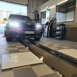 Szary SUV wjeżdża na stanowisko diagnostyczne z płytami pomiarowymi, widoczne oświetlenie warsztatowe i sprzęt diagnostyczny w tle.