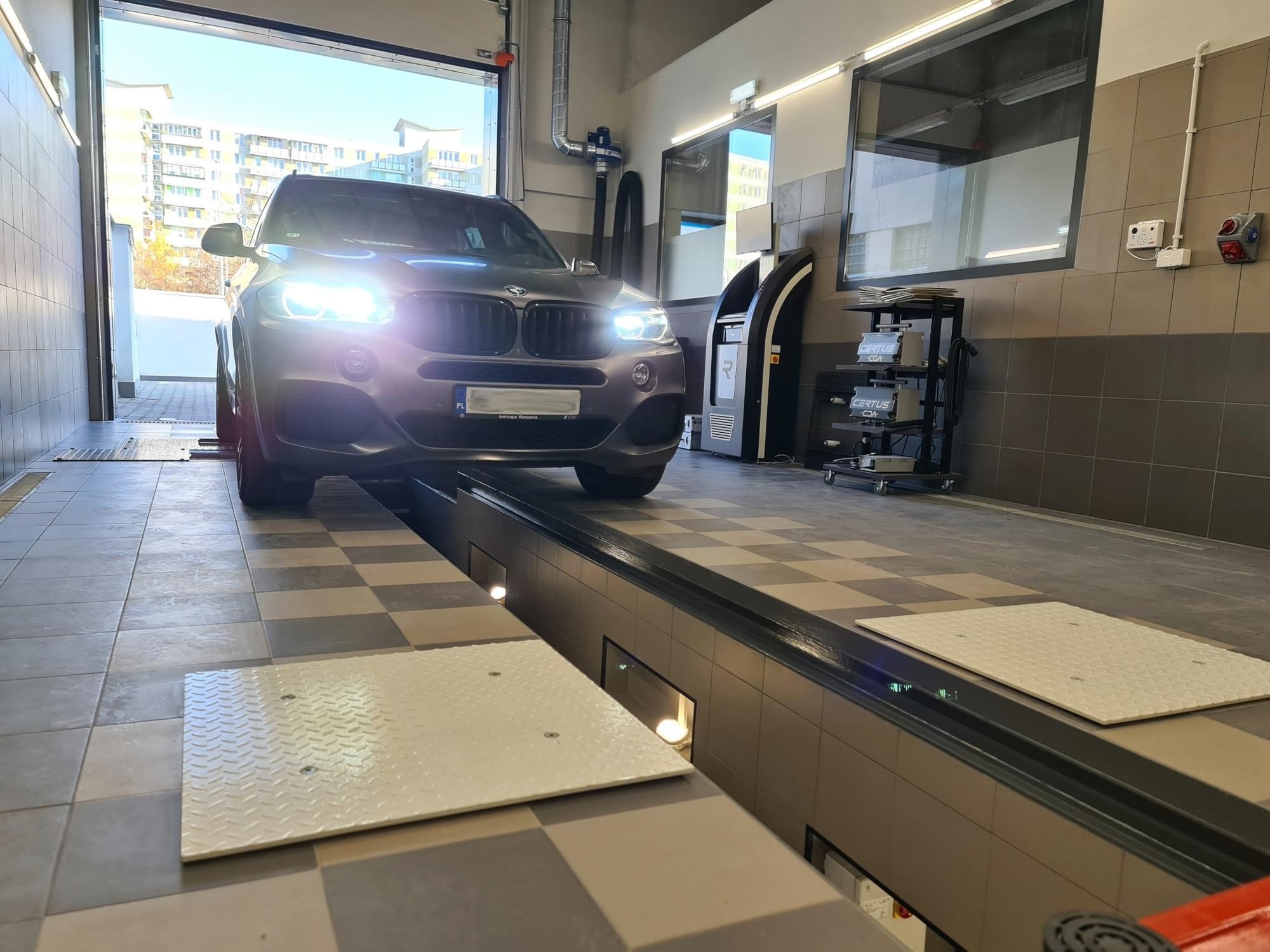 Szary SUV wjeżdża na stanowisko diagnostyczne z płytami pomiarowymi, widoczne oświetlenie warsztatowe i sprzęt diagnostyczny w tle.