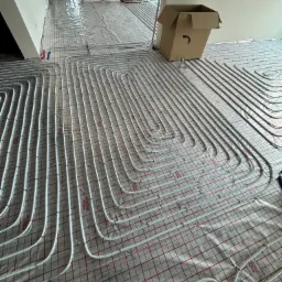 Układanie rur ogrzewania podłogowego na folii z siatką, widoczne złącza i mocowania, w tle pomieszczenie w trakcie remontu, kartonowe pudło.