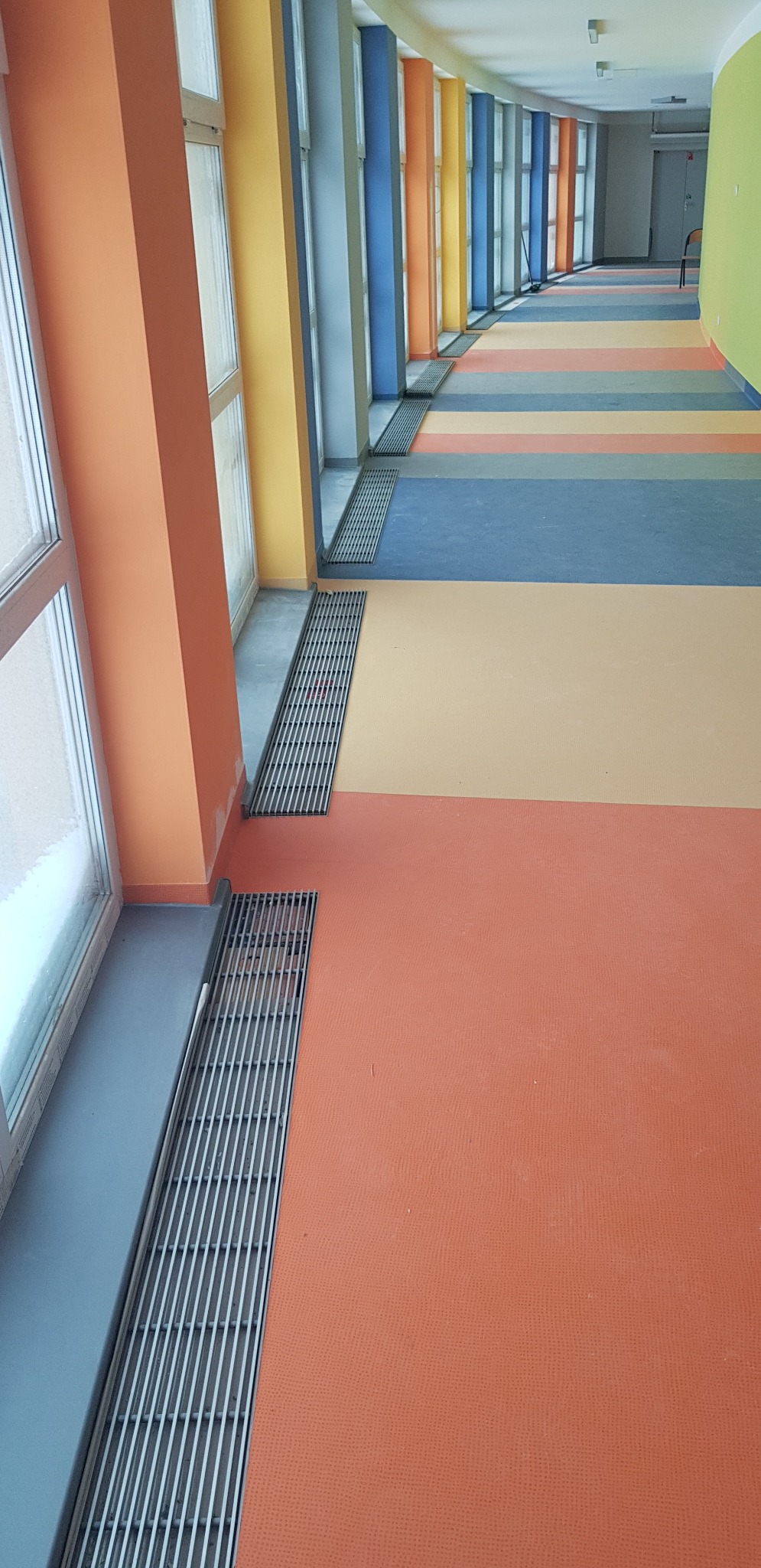 Długi korytarz z kolorowymi ścianami i podłogą w paski, widoczne kratki odpływowe wzdłuż okien.
