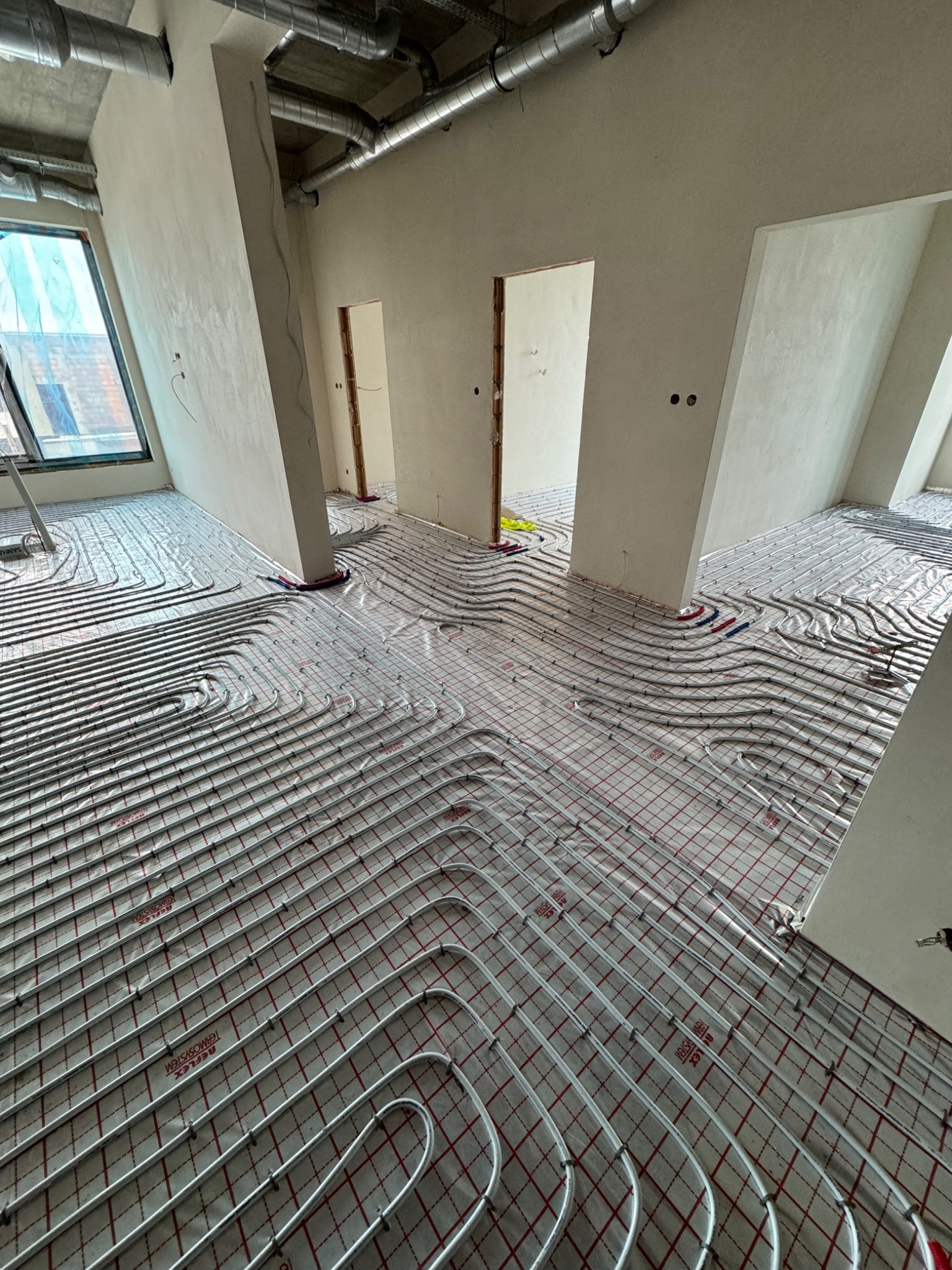 Białe rury ogrzewania podłogowego ułożone spiralnie na czerwonej siatce w surowym wnętrzu z widocznymi instalacjami wentylacyjnymi pod sufitem.