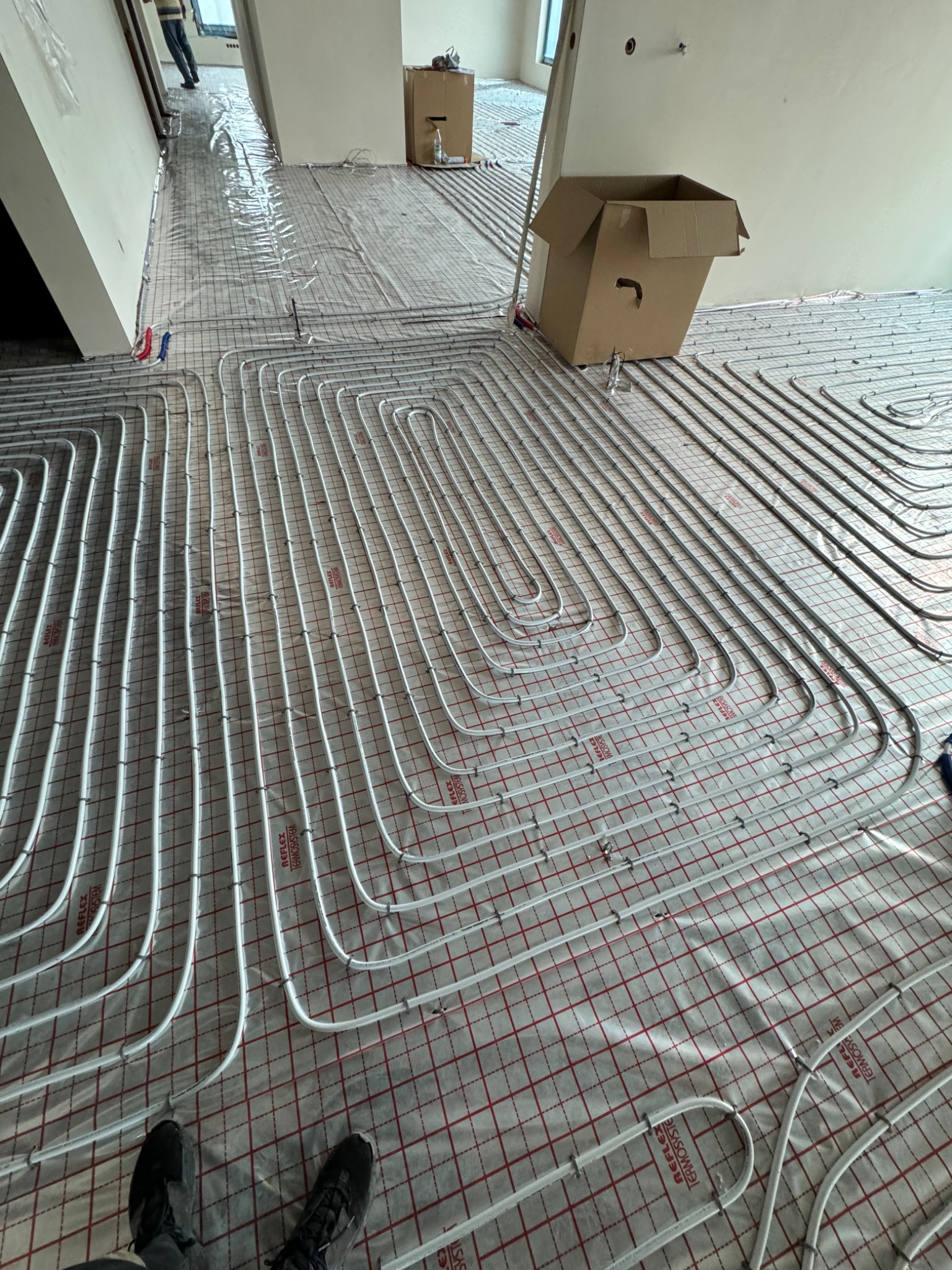 Układanie rur ogrzewania podłogowego na folii z siatką, widoczne złącza i mocowania, w tle pomieszczenie w trakcie remontu, kartonowe pudło.