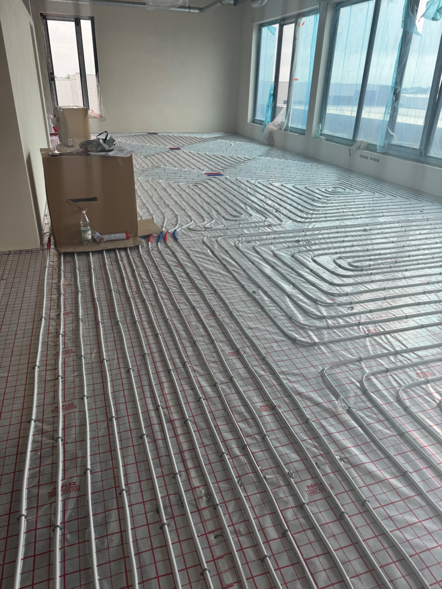 Białe rury ogrzewania podłogowego ułożone na folii z czerwoną siatką w przestronnym, jasnym pokoju z dużymi oknami; widoczny karton i butelka z płynem.