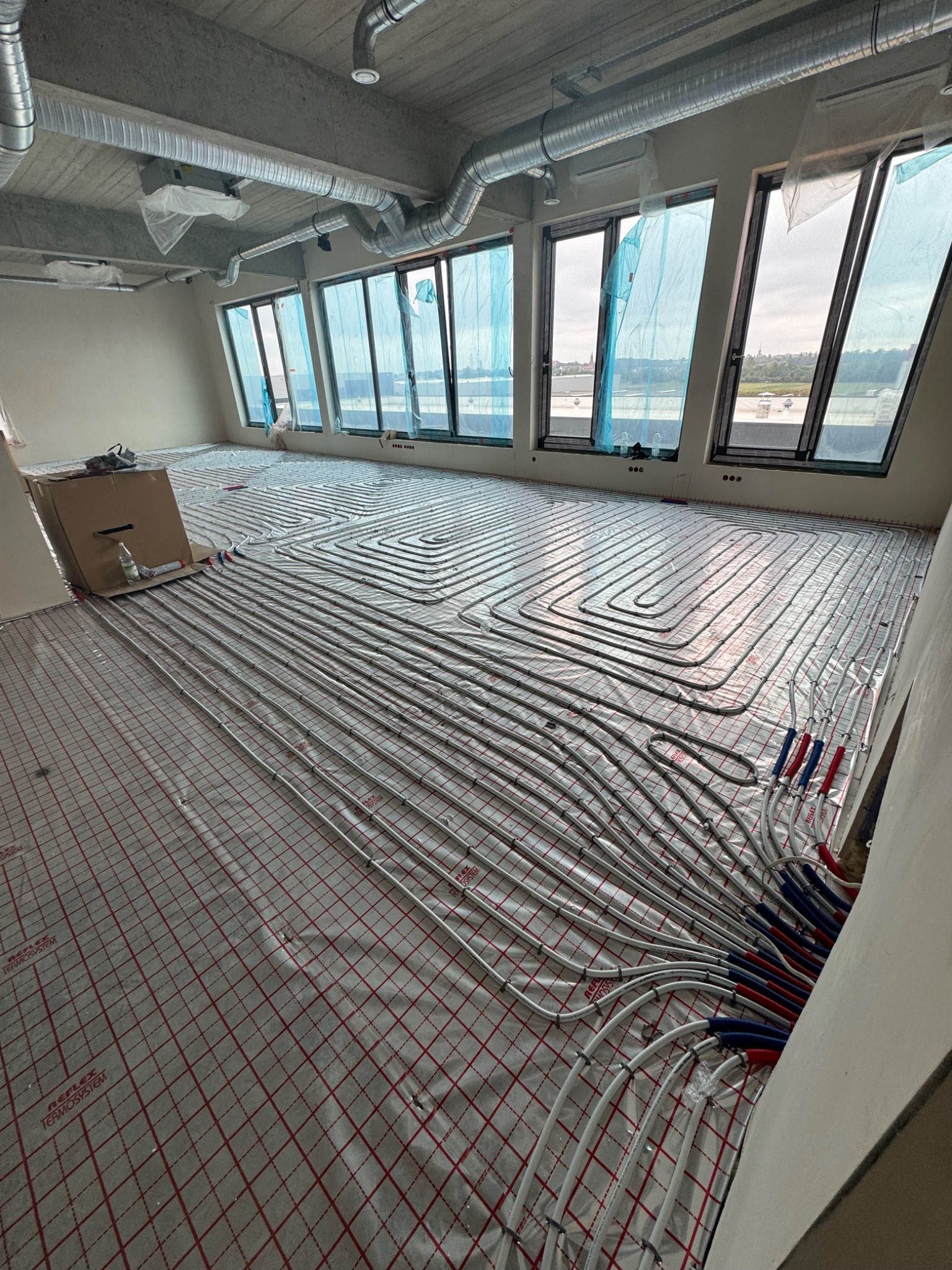 Rozłożone rury ogrzewania podłogowego na folii z siatką, widok na okna z niebieską folią zabezpieczającą i panoramą miasta w tle, instalacja w surowym stanie.