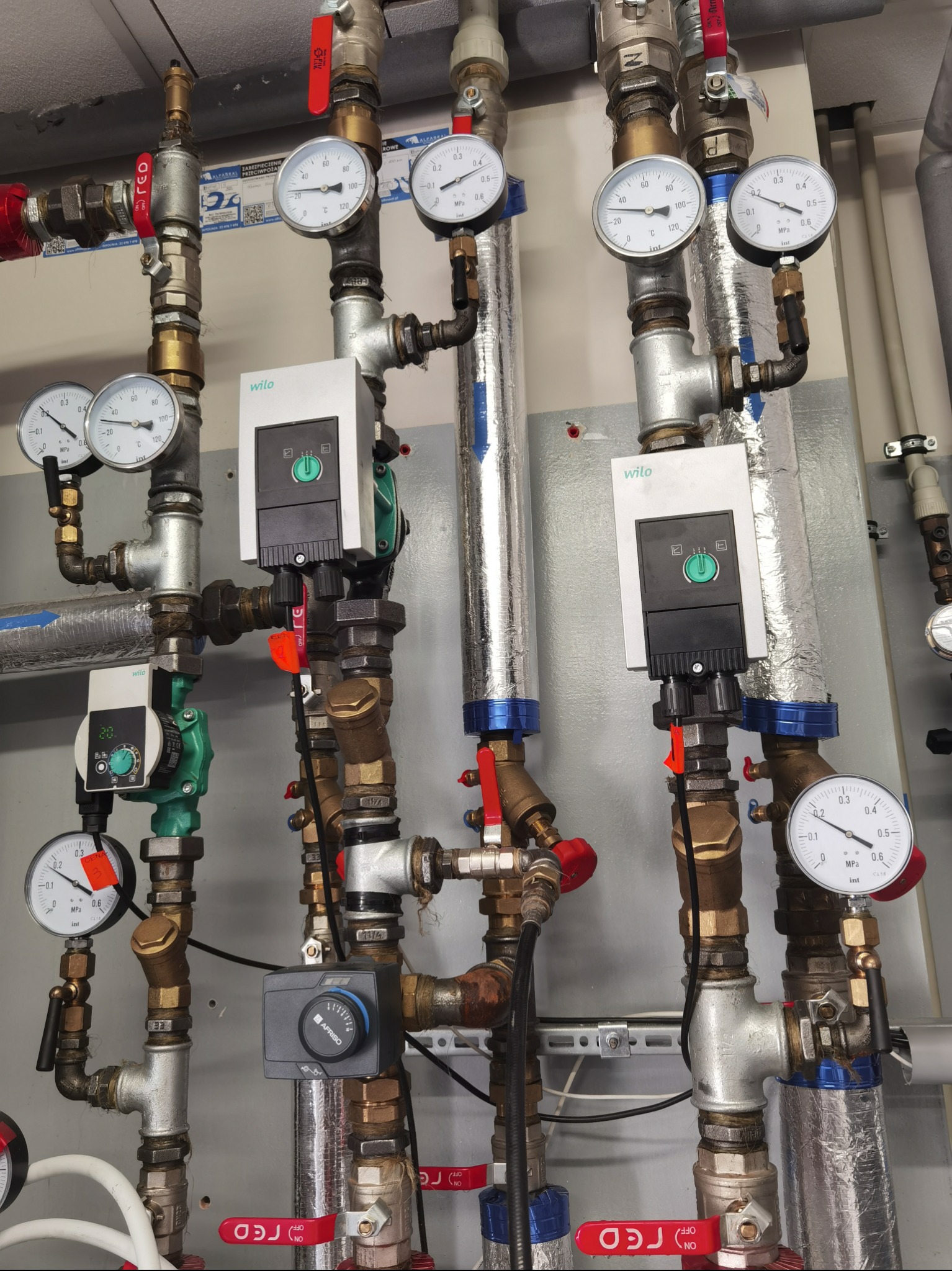Złożona instalacja hydrauliczna z rurami, zaworami, manometrami i sterownikami Wilo oraz Afriso, widoczne elementy zabezpieczenia przeciwpożarowego, całość na tle szarej ściany.