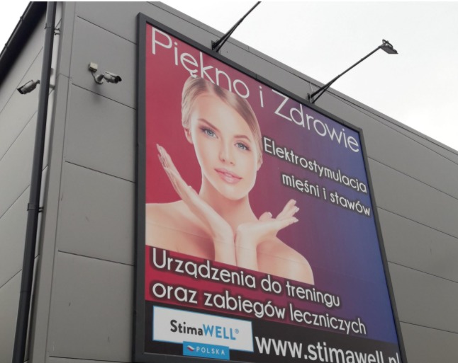 Zewnętrzny baner reklamowy na budynku z napisem 'Piękno i Zdrowie', prezentujący kobietę i informację o elektrostymulacji mięśni i stawów oraz urządzeniach do treningu i zabiegów leczniczych marki...