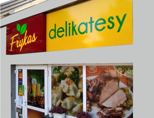 Fasada sklepu spożywczego 'Frykas' z szyldem w kolorach czerwonym i żółtym oraz oklejonymi witrynami prezentującymi pierogi, pieczeń i sałatkę jarzynową.