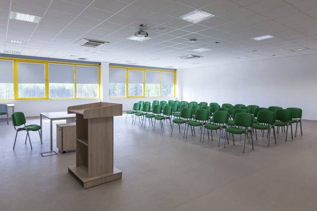 Sala konferencyjna z rzędem zielonych krzeseł, mównicą i oknami z widokiem na drzewa, oświetlona światłem dziennym i sztucznym.