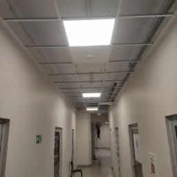 Korytarz biurowy z zamontowanym stelażem sufitu podwieszanego, widoczny pracownik na drabinie instalujący oświetlenie.