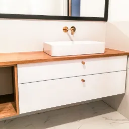 Nowoczesna łazienka z białą, prostokątną umywalką nablatową i złotą baterią ścienną, umieszczona na szafce z drewnianym blatem i białymi frontami szuflad z okrągłymi, złotymi uchwytami. Po lewej...