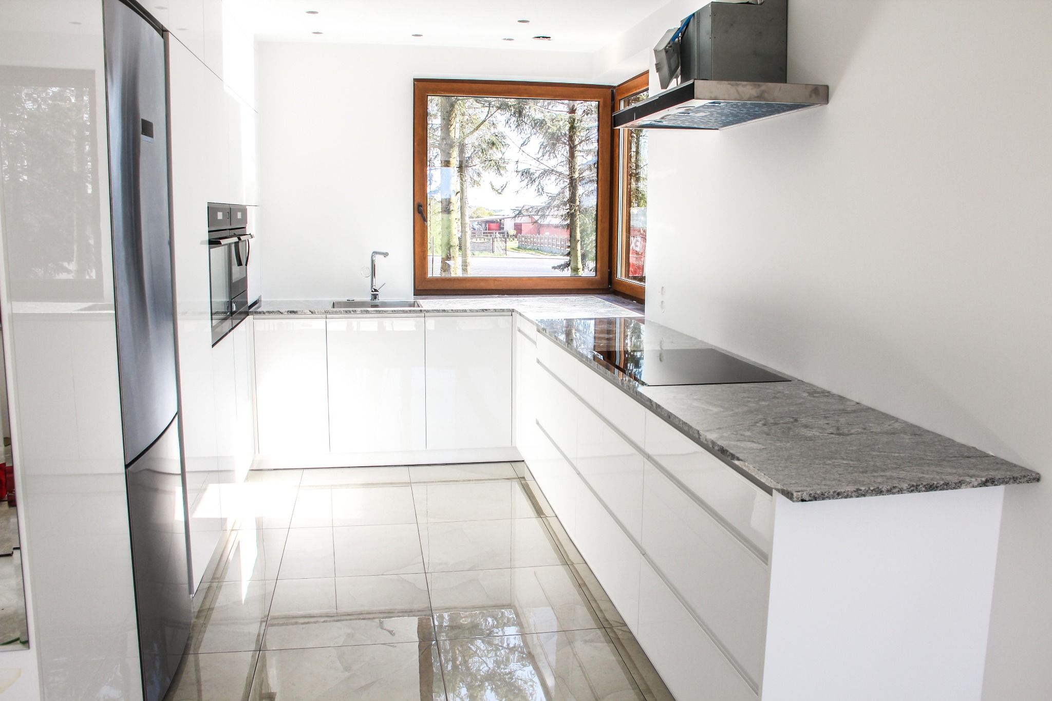 Jasna kuchnia z białymi szafkami, szarym blatem i oknem z widokiem na zieleń, błyszcząca podłoga z jasnych płytek odbija światło.